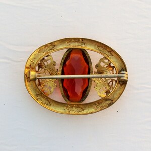 Op de afbeelding: Vintage ovale broche met een centrale gefacetteerde oranjerode steen. Het goudkleurige metalen frame heeft sierlijke filigraan en een sluiting met speld. Een klassiek sieraad.