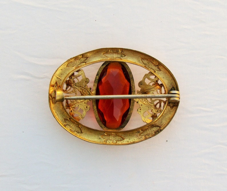 Op de afbeelding: Vintage ovale broche met een centrale gefacetteerde oranjerode steen. Het goudkleurige metalen frame heeft sierlijke filigraan en een sluiting met speld. Een klassiek sieraad.