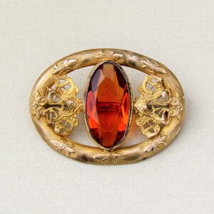 Op de afbeelding: Vintage, goudkleurige broche met een ovale, gefacetteerde, amberkleurige steen in het midden. De broche heeft een ovaal frame met sierlijke details in filigraanstijl en decoratieve accenten. Een klassiek sieraad.