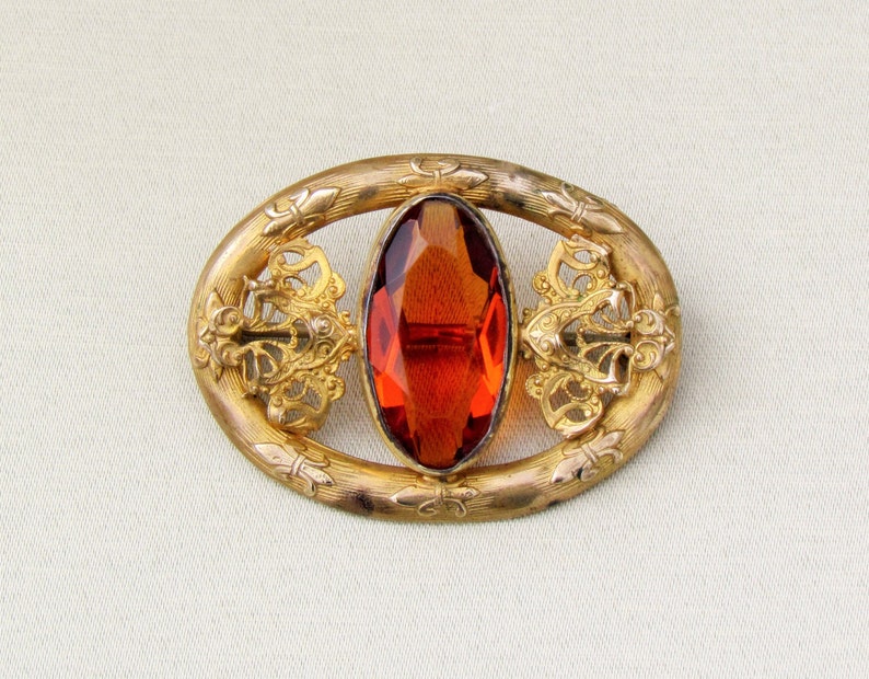 Op de afbeelding: Vintage, goudkleurige broche met een ovale, gefacetteerde, amberkleurige steen in het midden. De broche heeft een ovaal frame met sierlijke details in filigraanstijl en decoratieve accenten. Een klassiek sieraad.