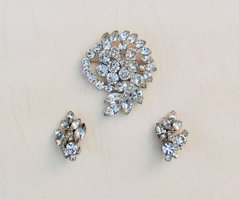 May include: A set of vintage rhinestone jewelry featuring a large brooch and a pair of earrings. The brooch is a swirl design with clear rhinestones and the earrings are smaller clusters of clear rhinestones.
