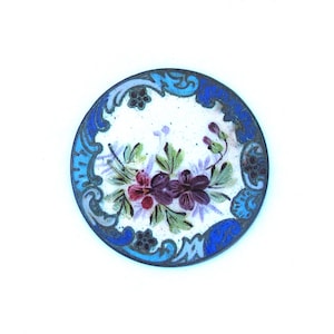 May include: A round, blue and white enamel button with a floral design. The button features a white background with a blue border and a floral design in shades of purple, pink, and green.