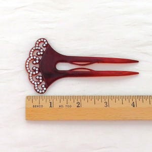 Puede incluir: Una horquilla para el cabello de color burdeos con un dise&ntilde;o de remolino decorativo adornado con pedrer&iacute;a transparente. El accesorio para el cabello mide aproximadamente 12.7 cm de largo y est&aacute; colocado junto a una regla de madera.