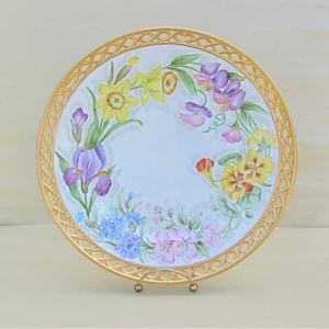 May include: A white porcelain plate with a gold rim and a woven design. The plate is decorated with hand-painted flowers in shades of yellow, purple, pink, and blue.