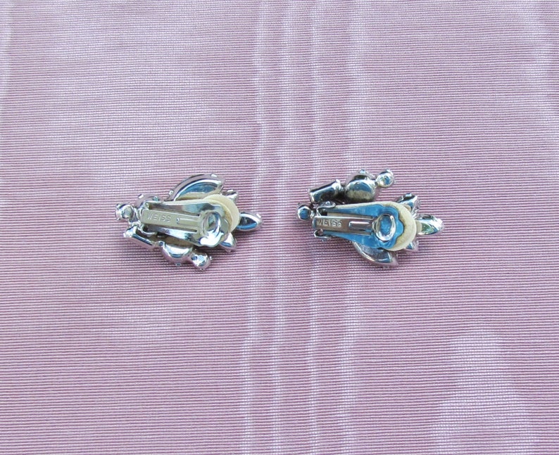 May include: A pair of silver tone clip-on earrings with clear rhinestones. The earrings have a floral design and the back of the earrings are marked "Weiss".