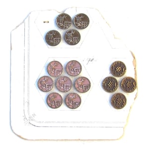 May include: A card with 15 vintage buttons. The buttons are arranged in two groups. The first group has 9 buttons with a pink background and a silver design of a deer. The second group has 6 buttons with a brown background and a gold design of a floral pattern. The card has the number "90" written on it.