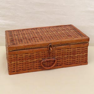 May include: A rectangular, woven wicker basket with a hinged lid. The basket is a warm brown color and features a handle on the front. The lid is secured with a metal clasp. This vintage-style basket is suitable for storage or display.
