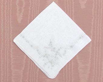 Op de afbeelding: Een witte zakdoek met delicate grijze bloemenborduurwerk in een hoek. De vierkante doek is gemaakt van een textuurstof en wordt gepresenteerd tegen een roze achtergrond. Een klassiek accessoire.