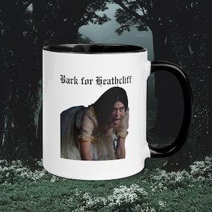 Może przedstawiać: Biały ceramiczny kubek z czarnym wnętrzem i uchwytem. Kubek zawiera napis "Bark for Heathcliff" gotycką czcionką, a także wizerunek osoby w sukni w stylu vintage. Kubek jest umieszczony na ciemnym tle, przypominającym las.
