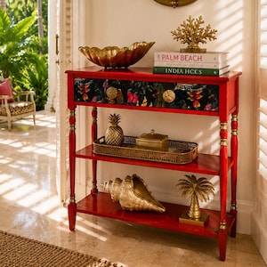 May include: A red console table with a floral drawer front, gold accents, and three tiers. It holds a gold shell bowl, a coral sculpture, books titled Palm Beach and India Hicks, a gold pineapple, a box, a large conch shell, and a palm tree figure.