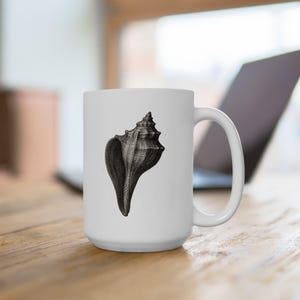 Könnte beinhalten: Weiße Keramiktasse mit einer detaillierten Schwarz-Weiß-Illustration einer Muschelschale. Die Tasse hat einen großen Henkel und steht vor einem verschwommenen Hintergrund mit einem Laptop und einer Holzoberfläche.