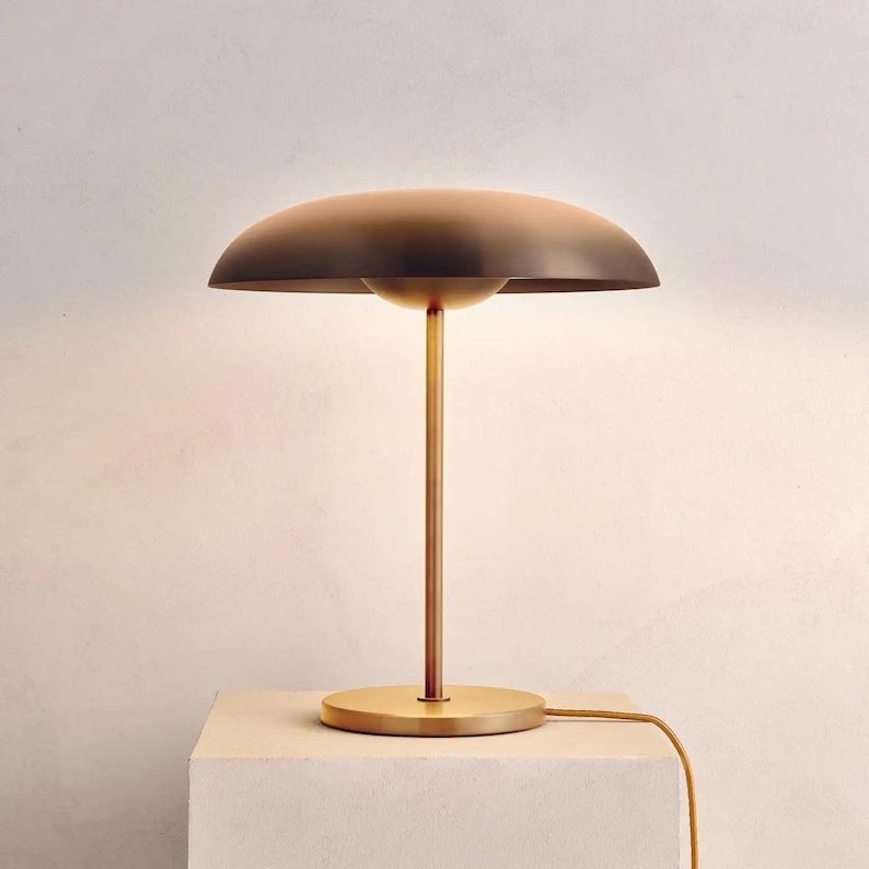 K&ouml;nnte beinhalten: Eine moderne Tischlampe aus Messing mit einem pilzf&ouml;rmigen Schirm. Die Lampe hat einen kreisf&ouml;rmigen Sockel, einen schlanken Stiel und einen gew&ouml;lbten Schirm. Der Schirm hat eine dunkelbraune Farbe, und die Lampe ist an eine Steckdose angeschlossen.