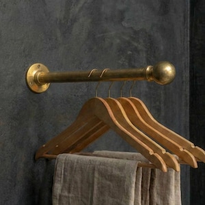 May include: A brass-coloured wall-mounted clothing rail with a round finial and mounting plate. Several wooden hangers are suspended from the rail, holding folded beige fabric. The rail is mounted on a dark grey textured wall.