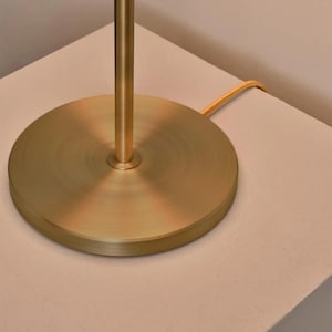 K&ouml;nnte beinhalten: Nahaufnahme eines goldfarbenen Lampenfu&szlig;es und -stiels. Der kreisf&ouml;rmige Sockel hat eine geb&uuml;rstete Oberfl&auml;che. Ein goldfarbenes Kabel erstreckt sich von der Lampe. Die Lampe steht auf einer hellen Oberfl&auml;che.