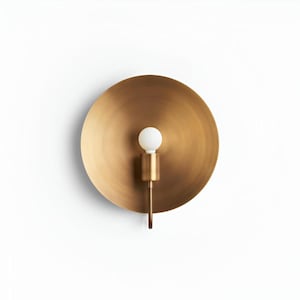 Può includere: Applique da parete in ottone con una grande piastra posteriore circolare e un piccolo apparecchio cilindrico. L'applique ha una finitura metallica calda e un design semplice e moderno. La lampadina è bianca.