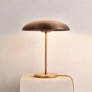 K&ouml;nnte beinhalten: Eine moderne Tischlampe aus Messing mit einem pilzf&ouml;rmigen Schirm. Die Lampe hat einen kreisf&ouml;rmigen Sockel, einen schlanken Stiel und einen gew&ouml;lbten Schirm. Der Schirm hat eine dunkelbraune Farbe, und die Lampe ist an eine Steckdose angeschlossen.