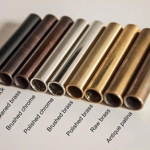 K&ouml;nnte beinhalten: Eine Auswahl an zylindrischen Metallr&ouml;hren in verschiedenen Ausf&uuml;hrungen, von Mattschwarz bis zu antiker Patina. Die R&ouml;hren sind mit Bezeichnungen wie 'Geb&uuml;rstetes Chrom' und 'Polierter Messing' versehen.