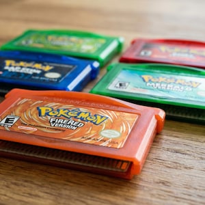 May include: An assortment of colorful Game Boy Advance cartridges, including an orange "Pokemon FireRed Version" cartridge. Other cartridges are green, blue, and red. The cartridges are arranged on a wooden surface.