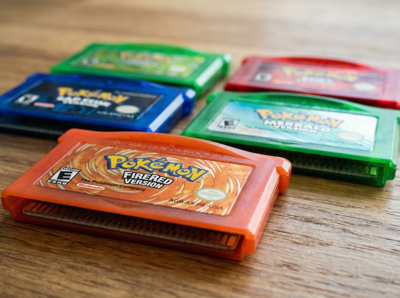 May include: An assortment of colorful Game Boy Advance cartridges, including an orange "Pokemon FireRed Version" cartridge. Other cartridges are green, blue, and red. The cartridges are arranged on a wooden surface.
