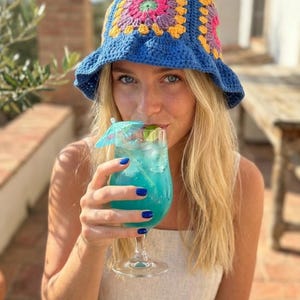 May include: A blue crochet bucket hat with a colorful square pattern on the crown. The hat has a wide brim. The person is holding a blue cocktail with a small umbrella. The person is wearing a white top.