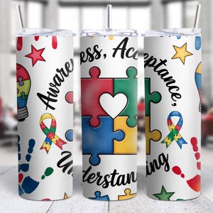 May include: White tumbler with a lid and straw, featuring colorful puzzle pieces, a heart, and a ribbon. The tumbler has the words "Awareness, Acceptance, Understanding" and various shapes and handprints.