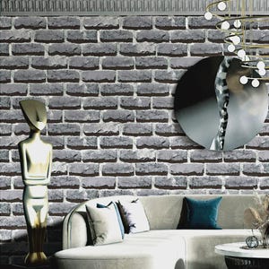 May include: A gray brick-patterned wallpaper covers the wall, with a decorative molding at the top. A gold-colored statue stands to the left of a white sofa with pillows. A round mirror and a modern chandelier are also visible.