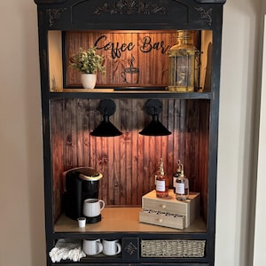 May include: Black coffee bar cabinet with a distressed finish. The cabinet features a coffee maker, mugs, and decorative items. The top shelf has the words "Coffee Bar" and a coffee cup logo. Two black sconces provide lighting.
