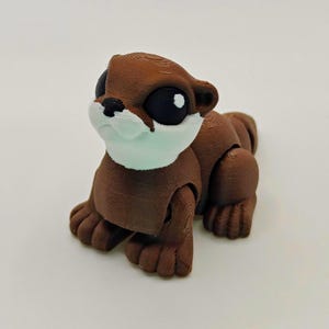 May include: A 3D-printed brown otter figurine with large black eyes and a white muzzle. The otter is in a seated position, with its paws and body detailed with textured lines. The figurine is a cute and whimsical animal-themed decoration.