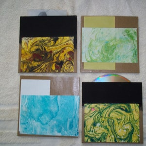May include: Four handmade CD cases with marbled paper covers. Each case has a black flap and a brown cardboard backing. The marbled paper designs feature yellow, green, blue, and red colors.