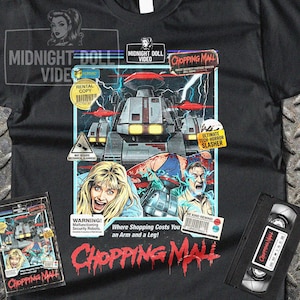 May include: Black t-shirt featuring a movie poster design for "Chopping Mall." The graphic includes a sci-fi horror scene with robots, distressed characters, and the film's title in red. Includes a VHS tape and movie cover.