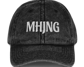 May include: A black acid-wash baseball cap featuring the white embroidered text MHJNG centered on the front panel. The cap has a six-panel construction with a curved brim, a top button, and visible stitching details across the crown and visor.