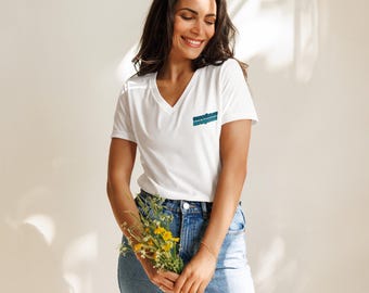 May include: A white V-neck t-shirt with the text "seashore magazine" in teal and blue. The shirt is paired with blue jeans. The person is holding a bouquet of yellow flowers.
