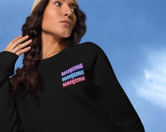May include: Black sweatshirt with the word "MAHJONG" stacked three times in a neon-style font. The sweatshirt is worn with a beige beanie. The background is a blue sky.