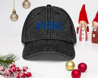 May include: A black acid-wash baseball cap featuring the text MHJNG embroidered in blue across the front panel. The cap has a structured crown, a curved brim, and stitched eyelets for ventilation, presenting a classic six-panel design.