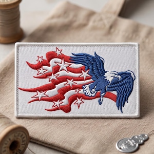 Op de afbeelding: Een rechthoekige geborduurde patch met een witte achtergrond, voorzien van een blauwe vliegende adelaar, vergezeld door rode golvende strepen en meerdere witte sterren. De patch ligt op een beige stoffen ondergrond bij twee houten garenklossen.