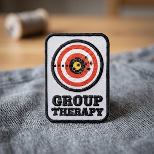 May include: A rectangular embroidered patch with a white background and a black border. The design features a circular red and white target with a yellow centre, marked with black bullet holes. The text GROUP THERAPY is stitched in black.