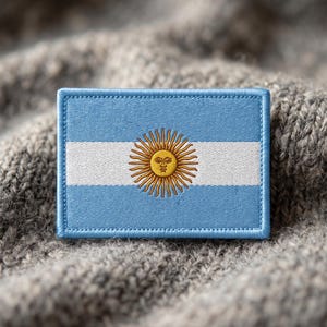 Op de afbeelding: Een rechthoekige geborduurde patch met de vlag van Argentinië. Het ontwerp bestaat uit drie horizontale banen in lichtblauw en wit, met een goudgeel zon-embleem met een menselijk gezicht in het midden van de witte baan.