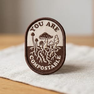 May include: An oval-shaped embroidered patch featuring a two-tone design of cream and dark brown. The patch displays a cluster of mushrooms with visible root systems and the text YOU ARE COMPOSTABLE embroidered in a dark brown thread.