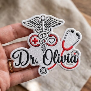 Può includere: Una toppa ricamata su sfondo bianco con cuciture nere e rosse. Presenta un simbolo del caduceo, una croce medica rossa, un cuore, uno stetoscopio e una piccola cuffia da infermiera. Il testo Dr. Olivia è ricamato in corsivo nero.