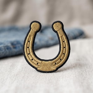 May include: A horseshoe-shaped embroidered patch featuring metallic gold thread detailing on a black fabric base. The design includes stitched nail hole accents along the curved frame, with a thick black border outlining the entire shape.