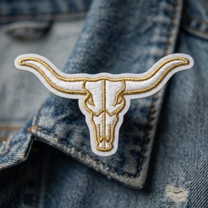 May include: An embroidered patch featuring a white longhorn steer skull design with metallic gold thread outlining the horns and facial structure. The patch is shaped to follow the contours of the skull and horns, creating a detailed graphic.