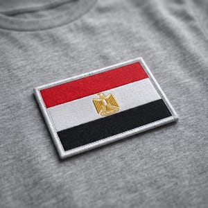 May include: A heather gray t-shirt featuring a rectangular embroidered patch of the Egyptian flag. The flag consists of horizontal red, white, and black stripes with a centered gold eagle emblem. The patch has a white stitched border.