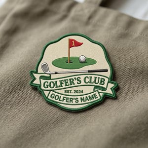 Könnte beinhalten: Ein kreisförmiger gestickter Aufnäher mit cremefarbenem Hintergrund und grünem Rand. Er zeigt einen Golfschläger, ein Green, einen weißen Golfball und eine rote Flagge mit der Zahl 1. Der Text lautet GOLFER'S CLUB, EST. 2024 und GOLFER'S NAME in Dunkelgrün.