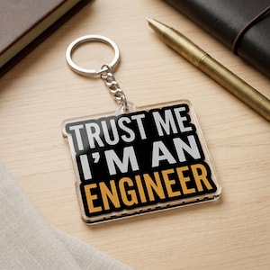 May include: A square acrylic keychain featuring a silver metal ring and chain. The keychain has a black background with the white text "TRUST ME I'M AN" and the yellow text "ENGINEER" printed in a bold, sans-serif font on the front surface.