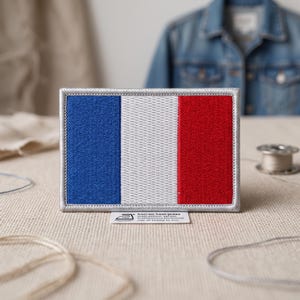 May include: A rectangular embroidered patch featuring the French flag with vertical stripes of blue, white, and red. The patch has a silver-grey border and a small white tag at the bottom with an iron icon and the text iron-on heat-press instructions.
