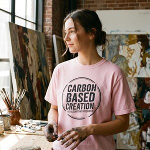 May include: A light pink t-shirt with the text "CARBON BASED CREATION" in a black circular design. The text "NO ALGORITHMS REQUIRED" is below the main text. The shirt is being worn by a person in an art studio.