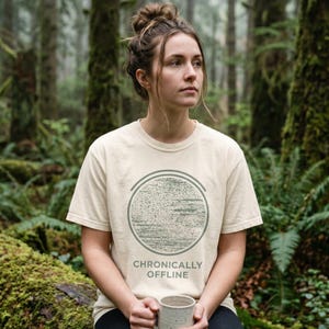 Op de afbeelding: Crèmekleurig T-shirt met een cirkelvormige afbeelding en de tekst "CHRONICALLY OFFLINE". De afbeelding is een textuurontwerp binnen twee concentrische cirkels. Het T-shirt heeft korte mouwen. De foto is in een bos gemaakt.