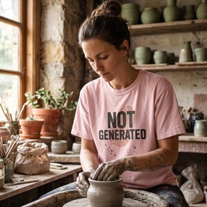 May include: A woman wearing a pink t-shirt with the words "NOT GENERATED" in black text is working on a pottery wheel. The pottery studio has shelves filled with green ceramic pieces and a window with potted plants.