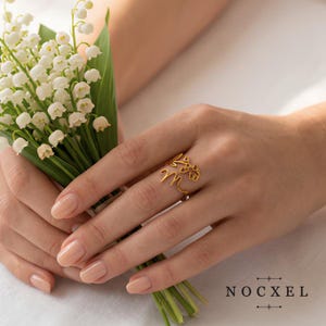 Peut inclure: Une bague dorée ornée d'une lettre M en écriture cursive et d'un petit motif floral, portée au doigt. La bague est ouverte et fabriquée en fil métallique fin. Le texte NOCXEL est imprimé dans le coin inférieur droit de l'image.