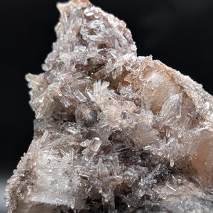 May include: Close-up of a mineral specimen featuring clusters of transparent, crystal-like formations. The crystals are a clear, icy color, with hints of brown and tan. The base of the specimen is a mix of gray and brown tones, creating a natural, earthy appearance.